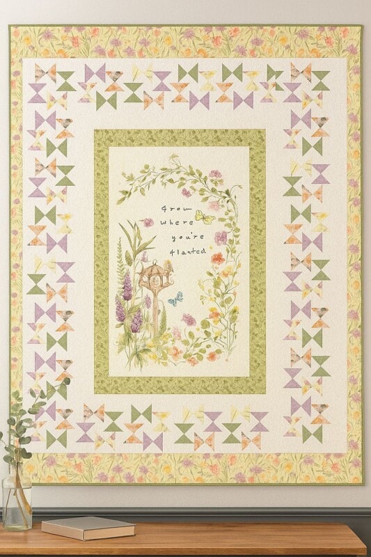 A decorative quilt hangs on a wall, featuring pastel pinwheel patterns, floral borders, and a central panel with flowers, a birdhouse, and the words “Grow where you’re planted.” Inspired by the Butterfly Garden Throw Quilt Pattern. A small vase with greenery sits below.