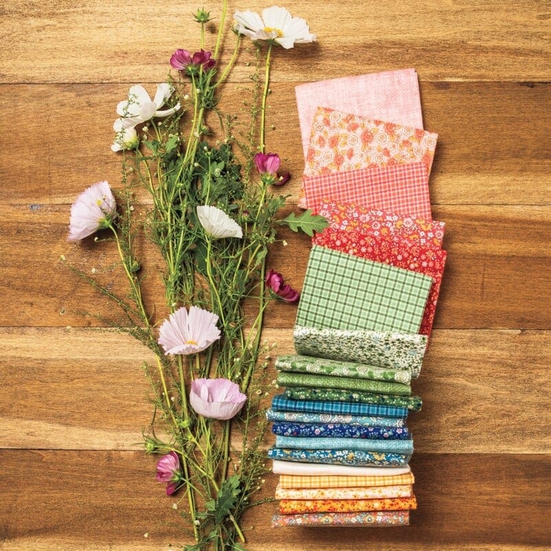 Colorful fabric squares in pink, red, green, blue, and yellow wholesale quilt patterns are stacked beside a bunch of delicate purple and white wildflowers, all arranged on a wooden surface.