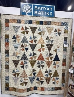 A multicolored patchwork quilt with assorted triangle patterns hangs on display under a “Banyan Batiks” sign at an indoor event or show. The quilt features earth-toned and blue batik fabrics.