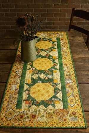 Sunflower-Award-on-table - A quilted table runner with yellow, green, and patterned patchwork designs lies on a rustic wooden table. A metal pitcher with dried flowers sits atop the runner, and a brick wall is in the background.