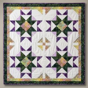 Casserole-Carry-on-wall A quilt with a symmetrical geometric pattern featuring green, purple, and pastel batik fabrics arranged in star shapes, framed by a dark green patterned border. The quilt hangs against a light-colored wall.