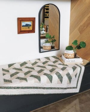 Rain-Chains Quilt A bench covered with a green and white patterned quilt sits below a curved wall mirror and a small framed landscape painting. Two potted plants and stacked books rest on the bench near the mirror.