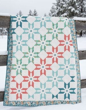 A colorful quilt with blue, teal, and red geometric patterns draped over a wooden fence, with snow covering the fence and a snowy landscape with trees in the background.