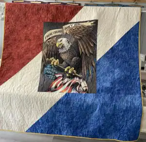 A quilt with red, white, and blue diagonal sections features a central image of a bald eagle grasping an American flag, with the words “These Colors Don’t Run” above the eagle.