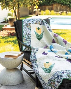 Gem-Dandy-Outside A colorful patchwork quilt with floral patterns draped over a black lounge chair beside a pool, with an open book resting on a small round stone table. Sunlight brightens the backyard scene.