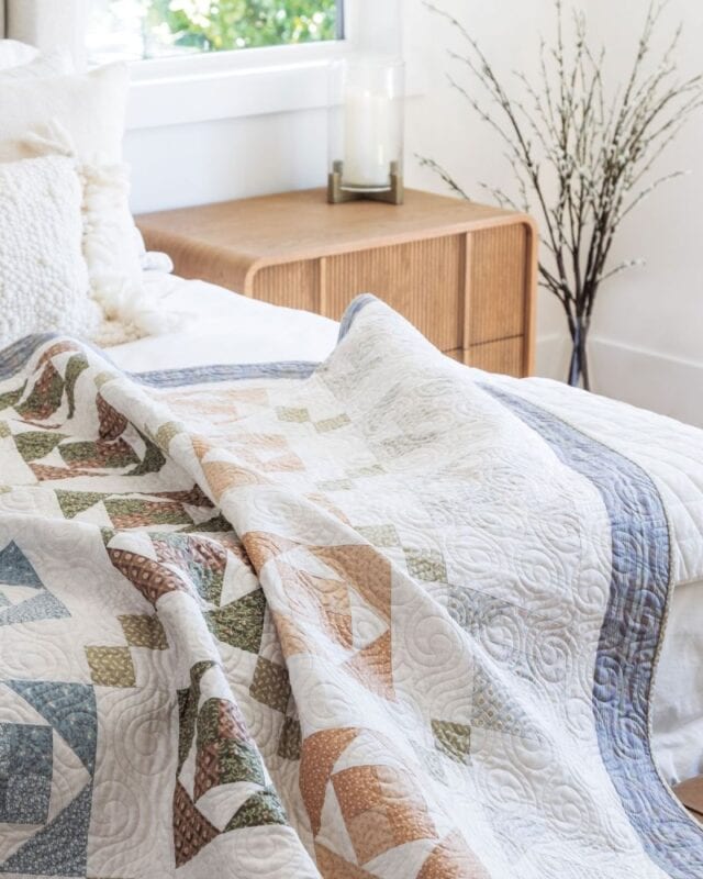 A colorful patchwork quilt with geometric patterns lies on a bed next to a wooden nightstand, a candle, and a vase with branches in a bright, cozy bedroom.