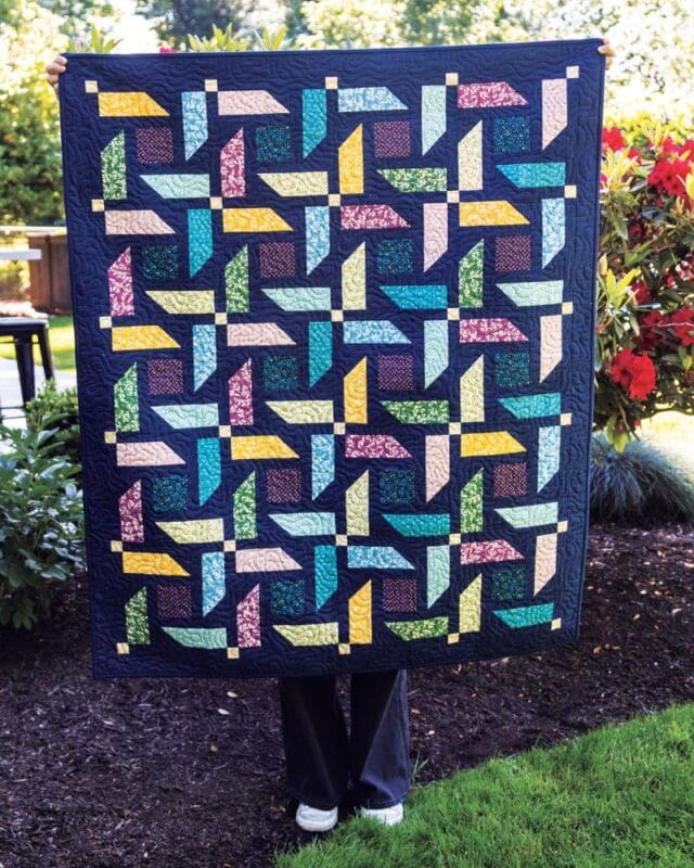A person stands outdoors, mostly hidden behind a colorful Turnabout Lap Quilt Pattern featuring geometric, pinwheel-like designs in blue, yellow, pink, and green. Lush green plants and red flowers bloom vibrantly in the background.