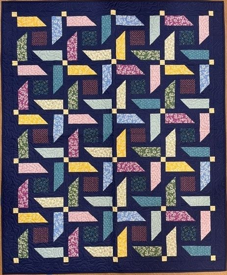A colorful Turnabout Lap Quilt Pattern featuring a geometric pinwheel design. Various fabric strips in pink, yellow, blue, green, lavender, and red create interlocking shapes on a dark blue background with small yellow squares.