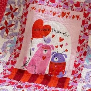A colorful quilt features two cartoon dogs, one pink and one purple, with hearts around them. A bird sits on the pink dog, holding a heart-shaped balloon that says welcome friends! above the characters.