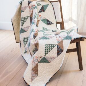 A colorful patchwork quilt with geometric patterns draped over a wooden chair on a light hardwood floor, with light curtains in the background.
