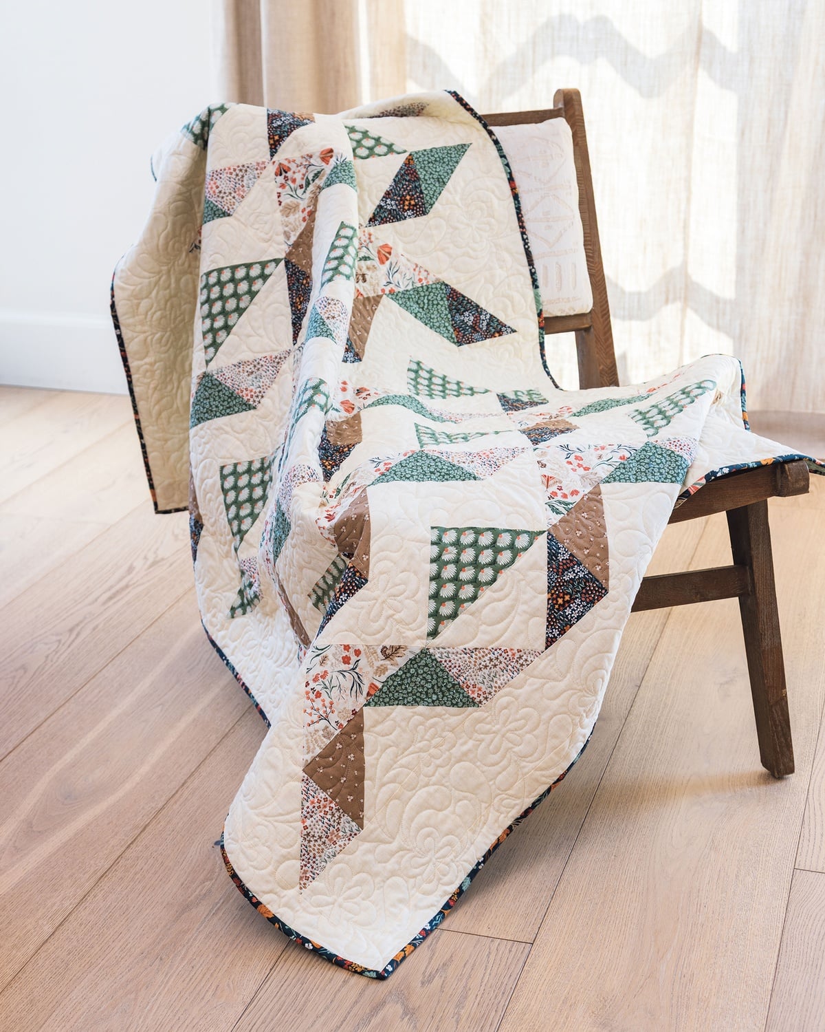A colorful patchwork quilt with geometric patterns draped over a wooden chair on a light hardwood floor, with light curtains in the background.