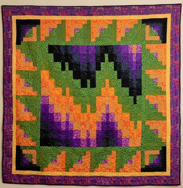 A colorful quilt features a geometric pattern with green, orange, purple, and black shapes reminiscent of a classic Log Cabin Bargello Quilt Pattern. The border has purple and yellow tones with patterns and symbols, enhancing the artistic appeal. The quilt boasts a grid-like design with zigzag and triangular motifs throughout.