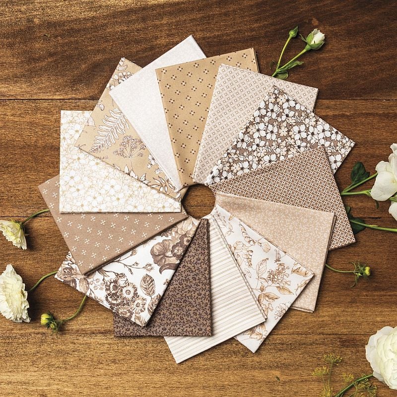 Crossroads Table Runner fabrics, a fan of cream, tan, and brown fat quarters on a wood table with white roses