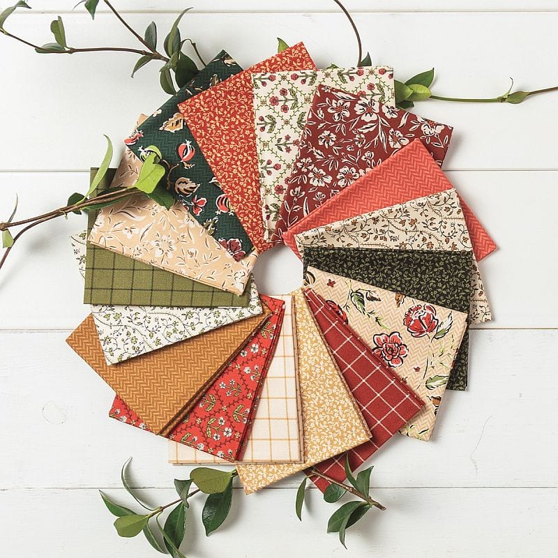 Fat quarter fan of red, green, caramel, and cream fabrics for the Cider and Cloves quilt, with green vine sprigs